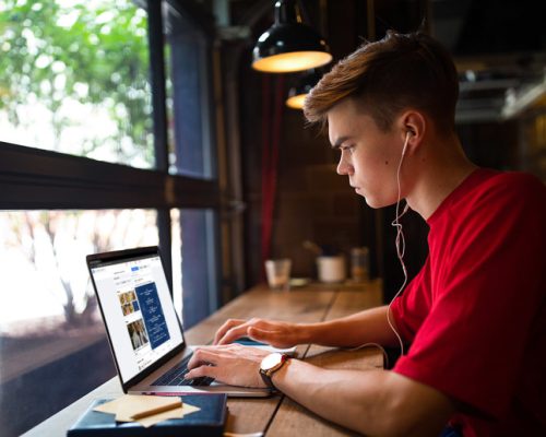 Hipster guy in casual wear listening to music via headphones and online learning via laptop computer while relaxing in coffee shop. Man skilled blog writer typing text on notebook gadget